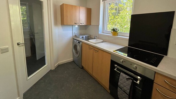 A compact kitchen with a washing machine, a sink, a stove, and wooden cabinets. - Two Bedroom Cottage in Lumsden - Contractors (Huntly)