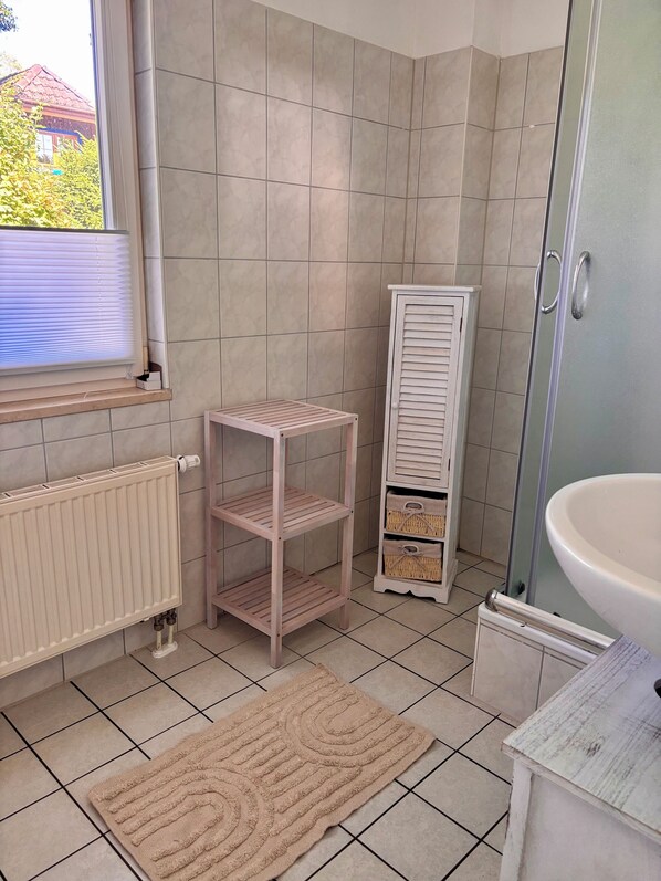 Ein Badezimmer mit gefliestem Boden, einem Heizkörper, einem Regal und einem Korb-Aufbewahrungsmodul sowie einem Waschbecken.