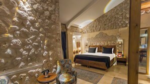 Une chambre avec un mur en pierre, un lit doté d’une tête de lit sombre, un fauteuil à motifs floraux et une petite table.