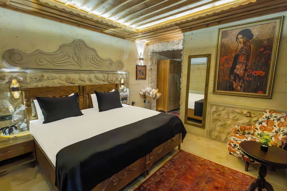 A bedroom with a large bed, a nightstand, a floral armchair, a small table with a vase, and a painting on the wall.
