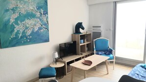 A modern living room with a large painting, a flat-screen TV, a bookshelf, a blue chair, a small table, and a sofa. - Exceptional view of the sea from the popular Corniche district (Sète)