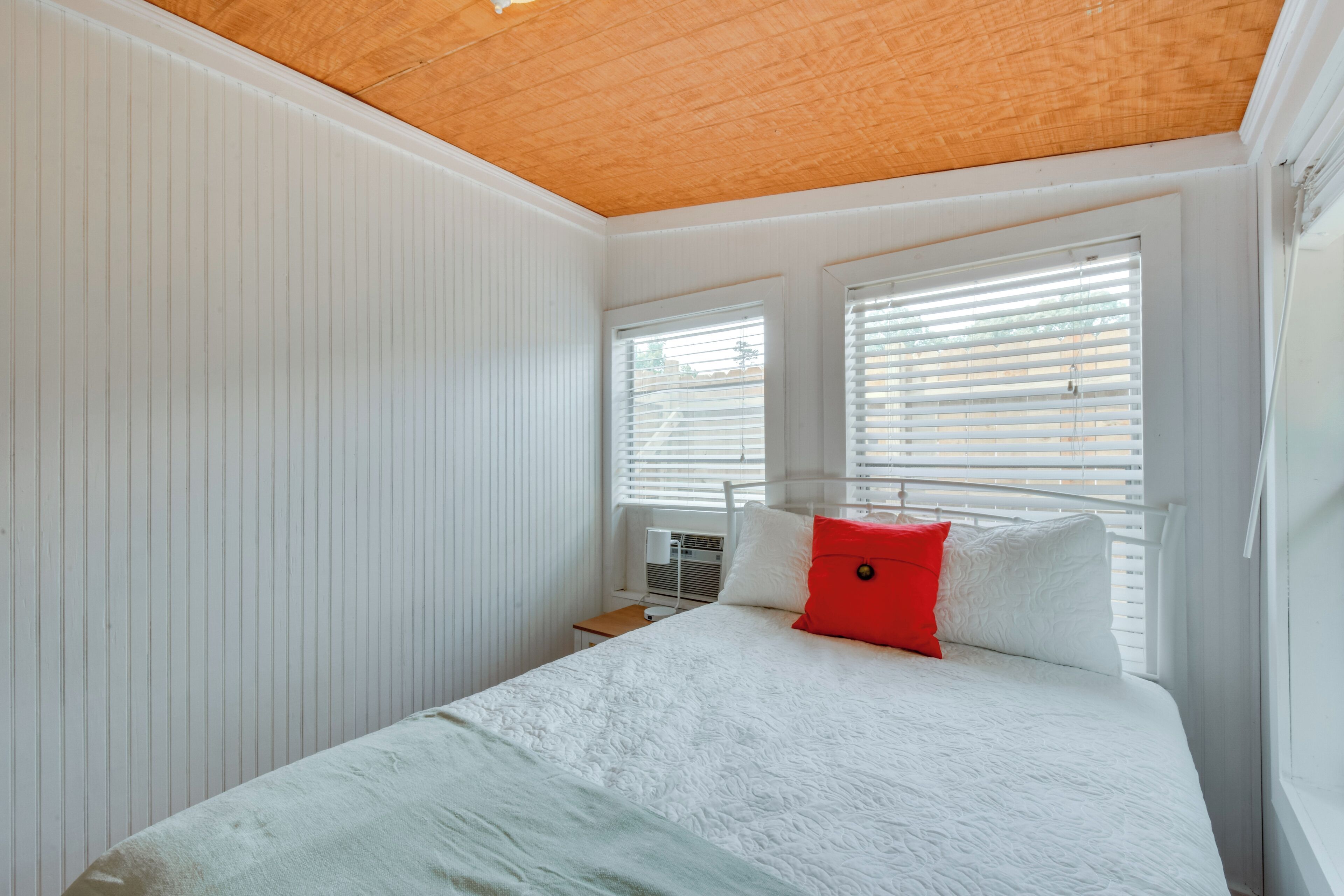 Un dormitorio con techo de madera, paredes blancas, una cama con ropa de cama blanca y roja, y una ventana con persianas.