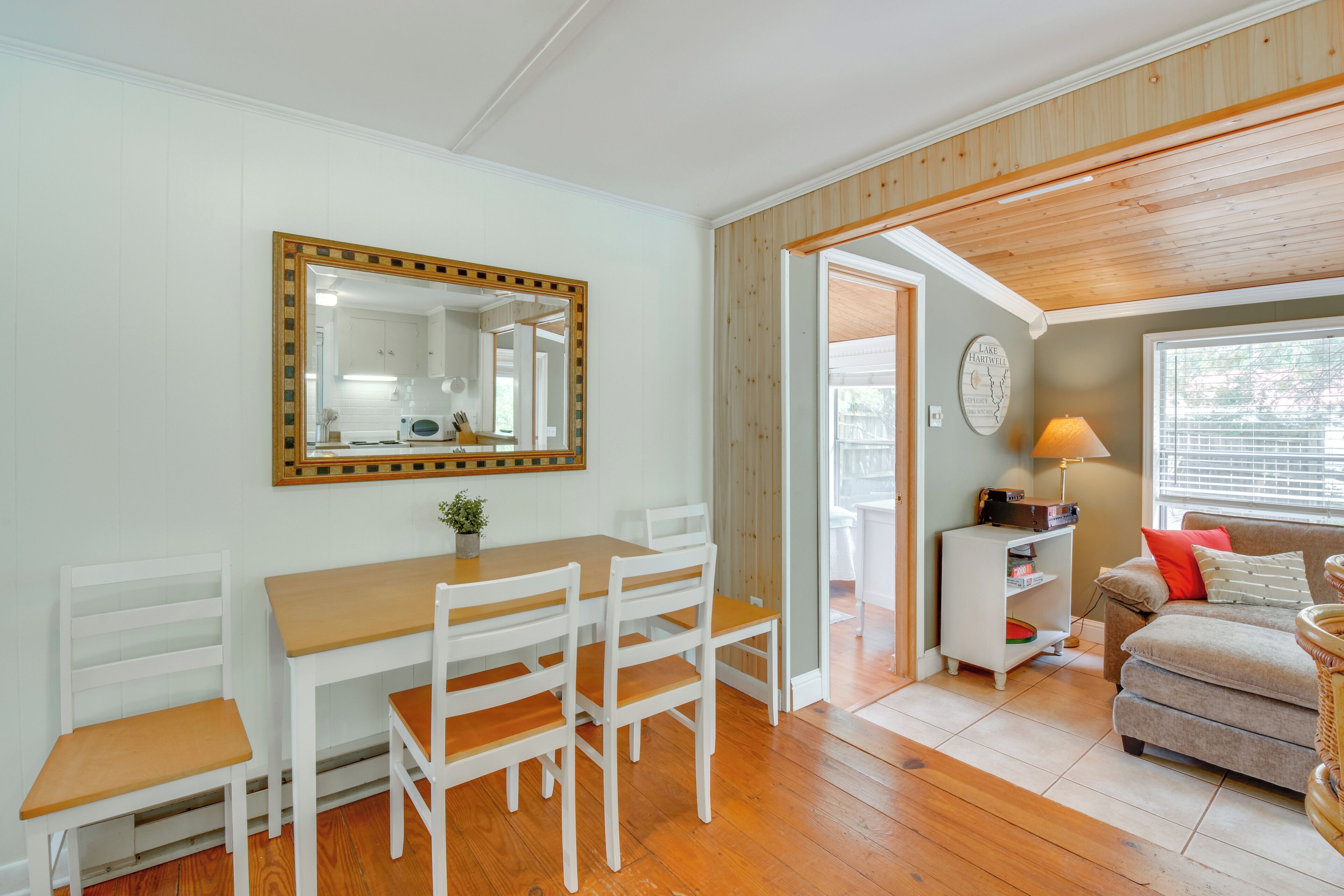 A dining area with a wooden table and white chairs, a mirror with a decorative frame, and a kitchen visible through an open door.