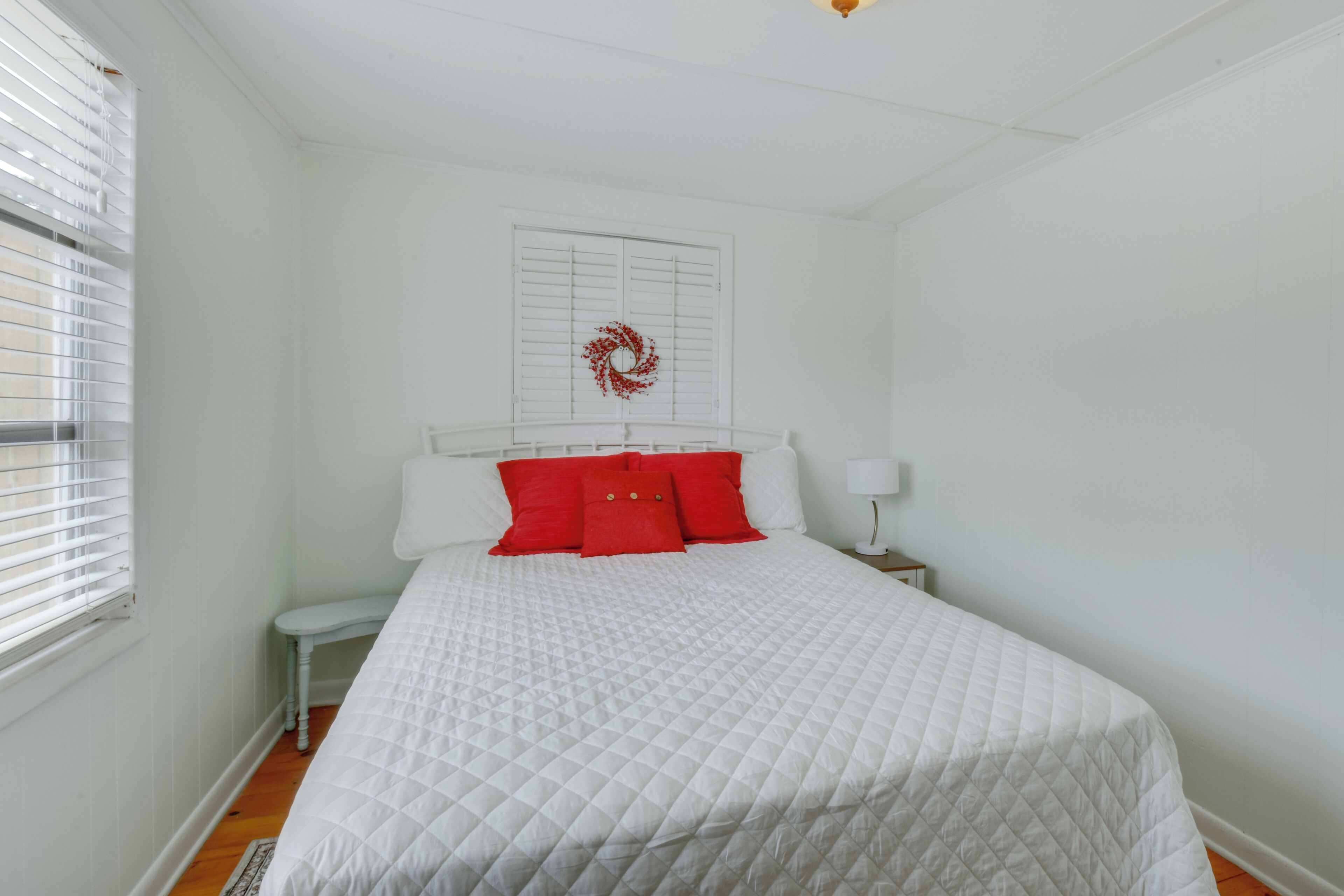 A bedroom with a white bed, red pillows, a white nightstand, and a white wall with a wreath.