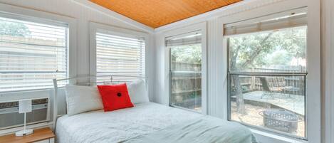 A bedroom with a bed, a nightstand, a window with blinds, and a view of an outdoor patio.