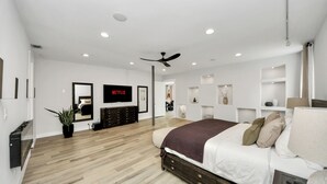 Un dormitorio moderno con una cama grande, ventilador de techo, iluminación empotrada, televisor y estanterías integradas.