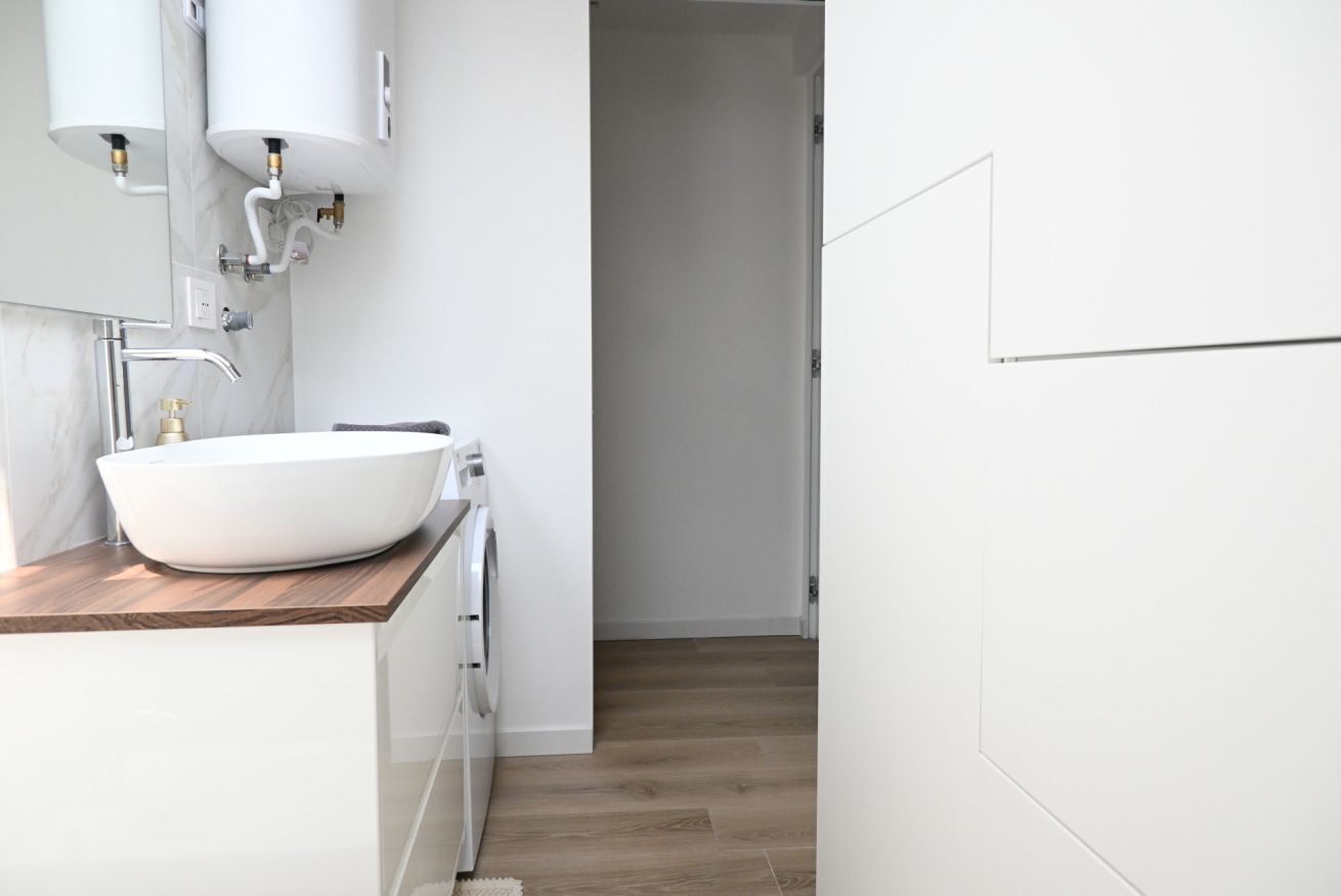 Un baño moderno con un lavabo blanco, encimera de madera y zona de lavandería a la vista.