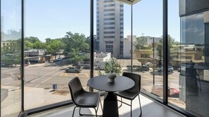 Ein Zimmer mit großem Fenster, das auf einen Parkplatz und Gebäude blickt. Ein runder Tisch mit zwei Stühlen und einer Vase steht darin.