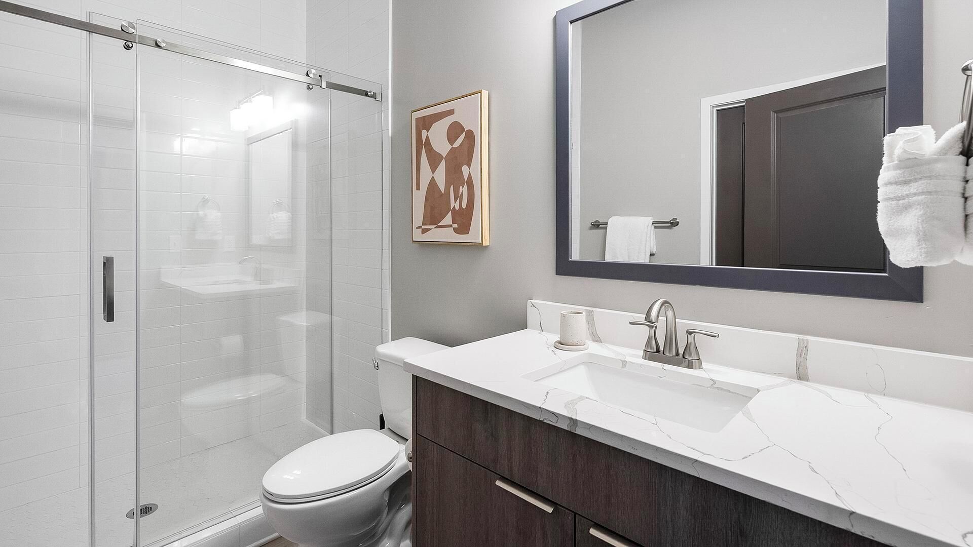 Un baño moderno con cabina de ducha de vidrio, inodoro blanco, lavabo de mármol y un espejo grande.