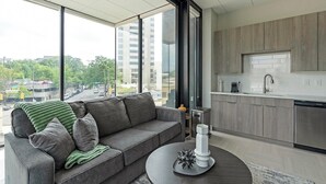 A modern living room with a grey sofa, a round coffee table, and a view of a city street through large windows.
