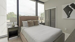 A modern hotel room with a large bed, two bedside tables with lamps, a wardrobe, and a view of the cityscape through large windows.