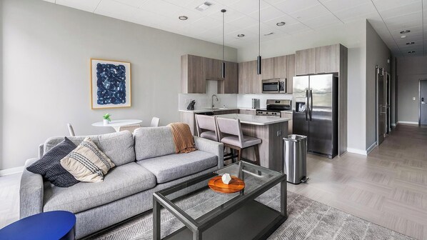 Una sala de estar moderna con un sofá gris, una mesa de centro y una zona de cocina con electrodomésticos de acero inoxidable.