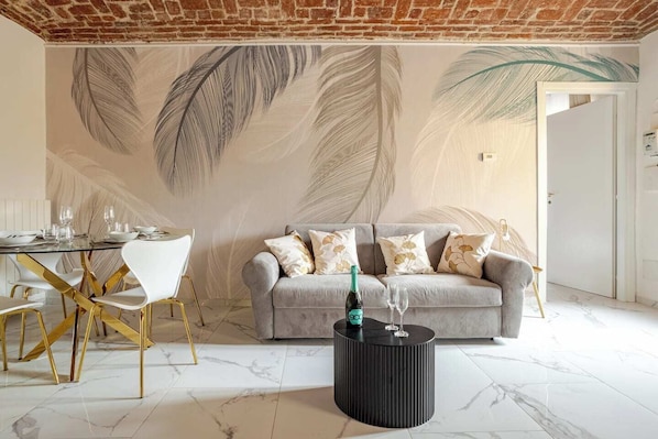Un salon moderne avec un canapé gris, des chaises blanches, une table basse ronde noire et une fresque murale représentant des motifs de plumes.