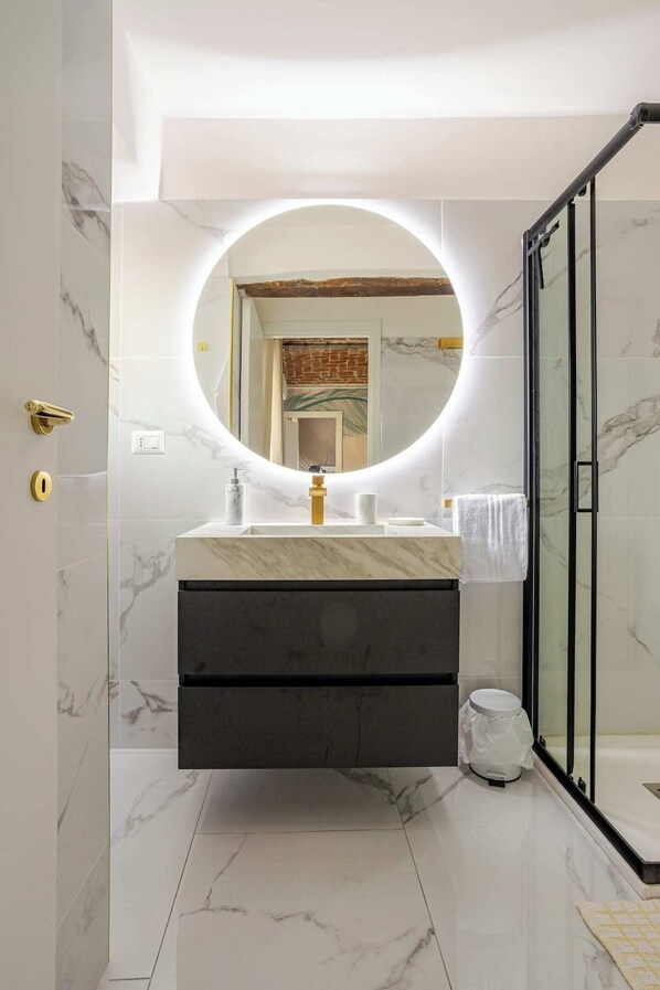 A modern bathroom with a marble floor, a round mirror with a light, a dark vanity, and a glass shower enclosure.