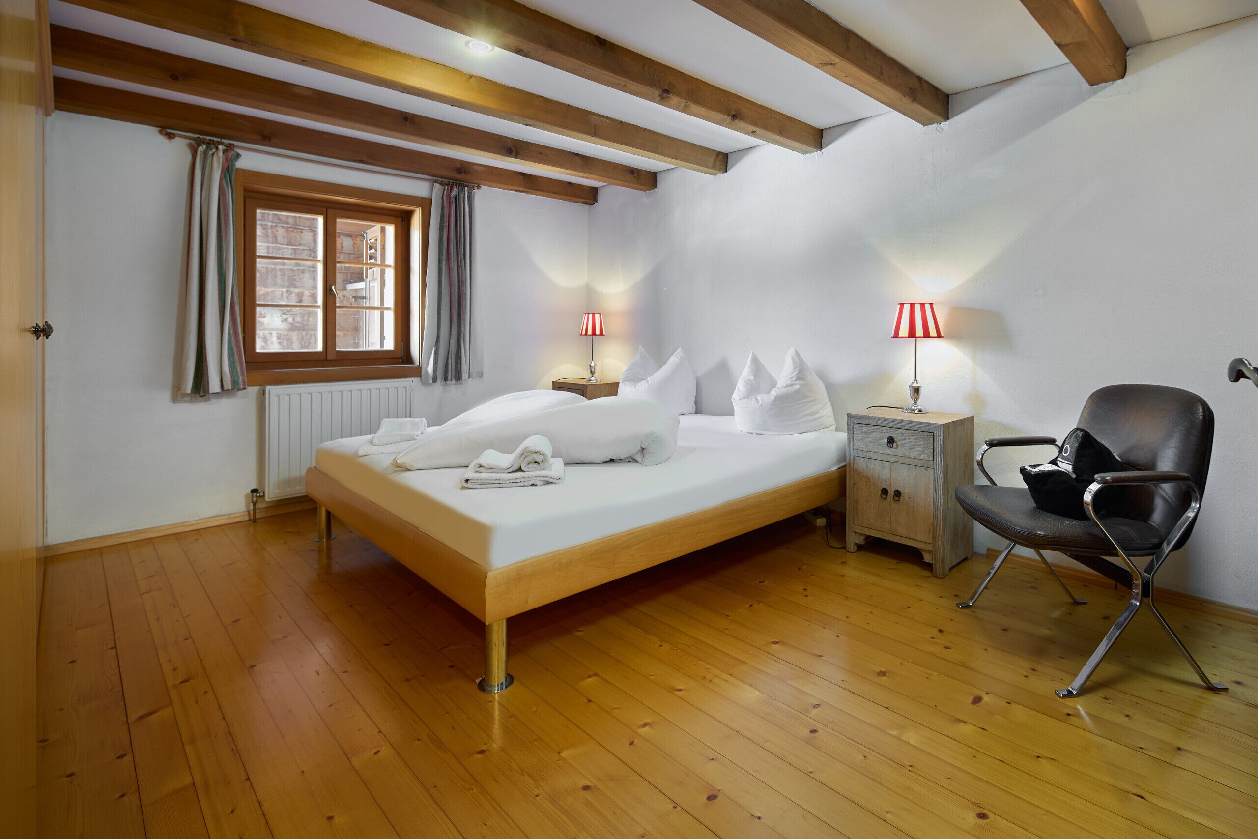 Ein Schlafzimmer mit einem hölzernen Bett, zwei Nachttischen mit Lampen, einem Sessel und einem Fenster mit Vorhängen.