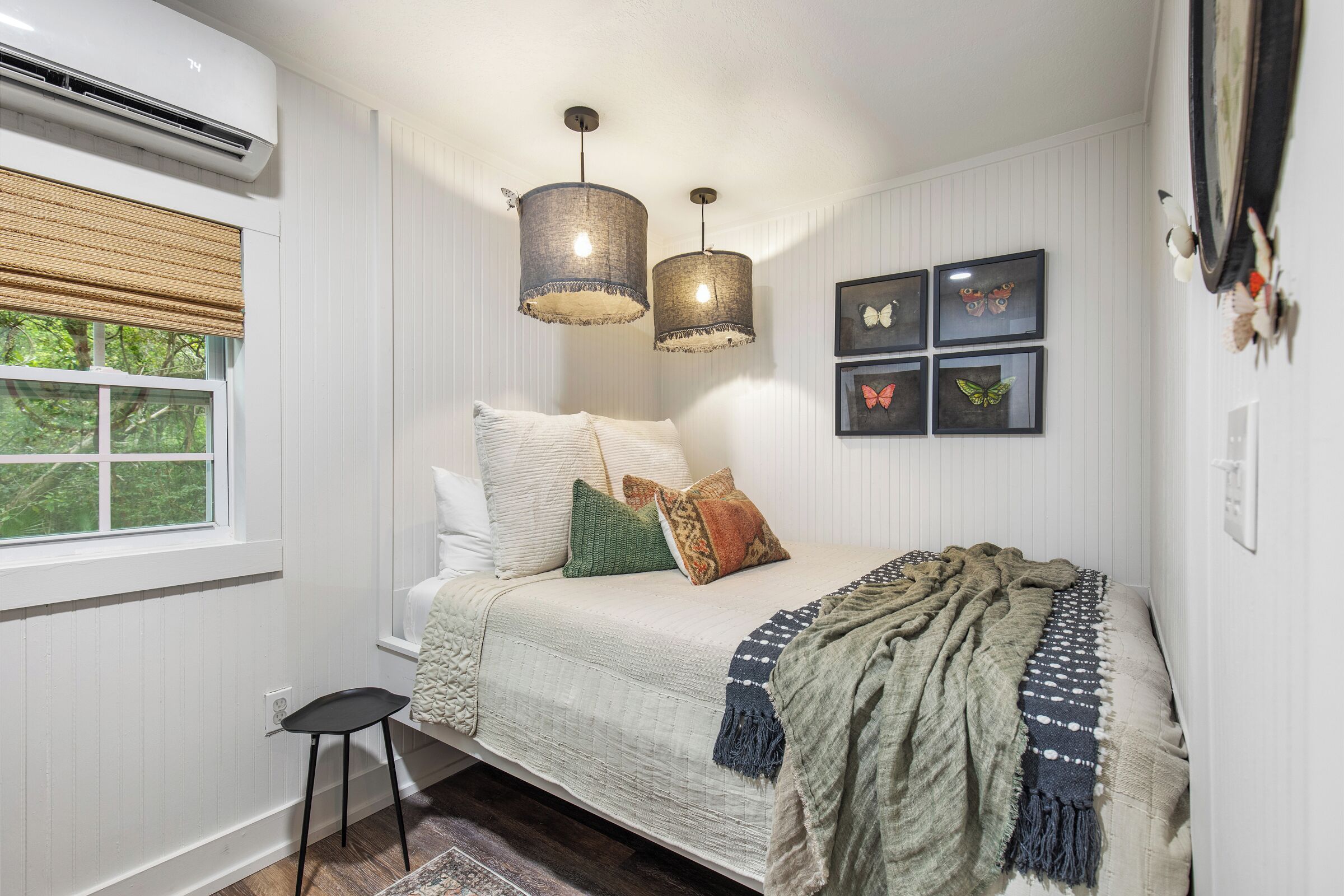 A bedroom with a bed, two hanging lamps, a window with blinds, and wall-mounted butterfly art.
