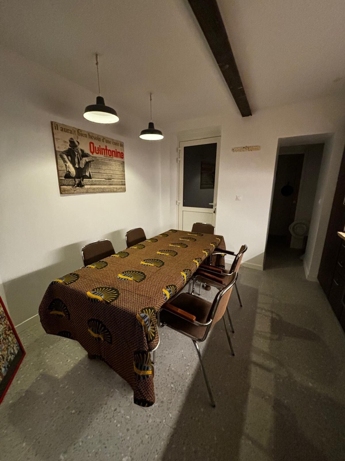 Un espace repas avec une longue table, des chaises, des luminaires suspendus et une affiche accrochée au mur.