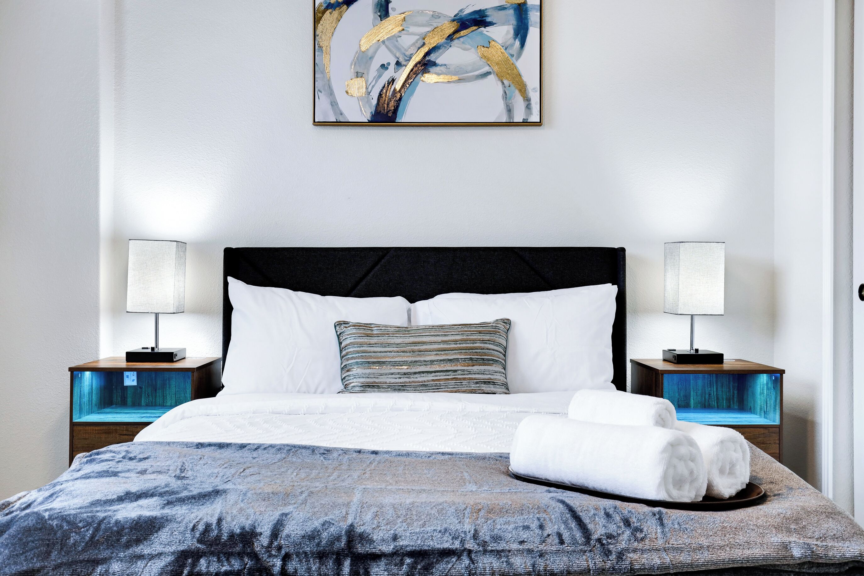Ein ordentlich gemachtes Bett mit weißen Bettwäsche, einem blauen Kissen mit Muster und einem einzelnen Streifen-Kissen. Auf jedem Nachttisch stehen Lampen und ein kleines Aquarium.