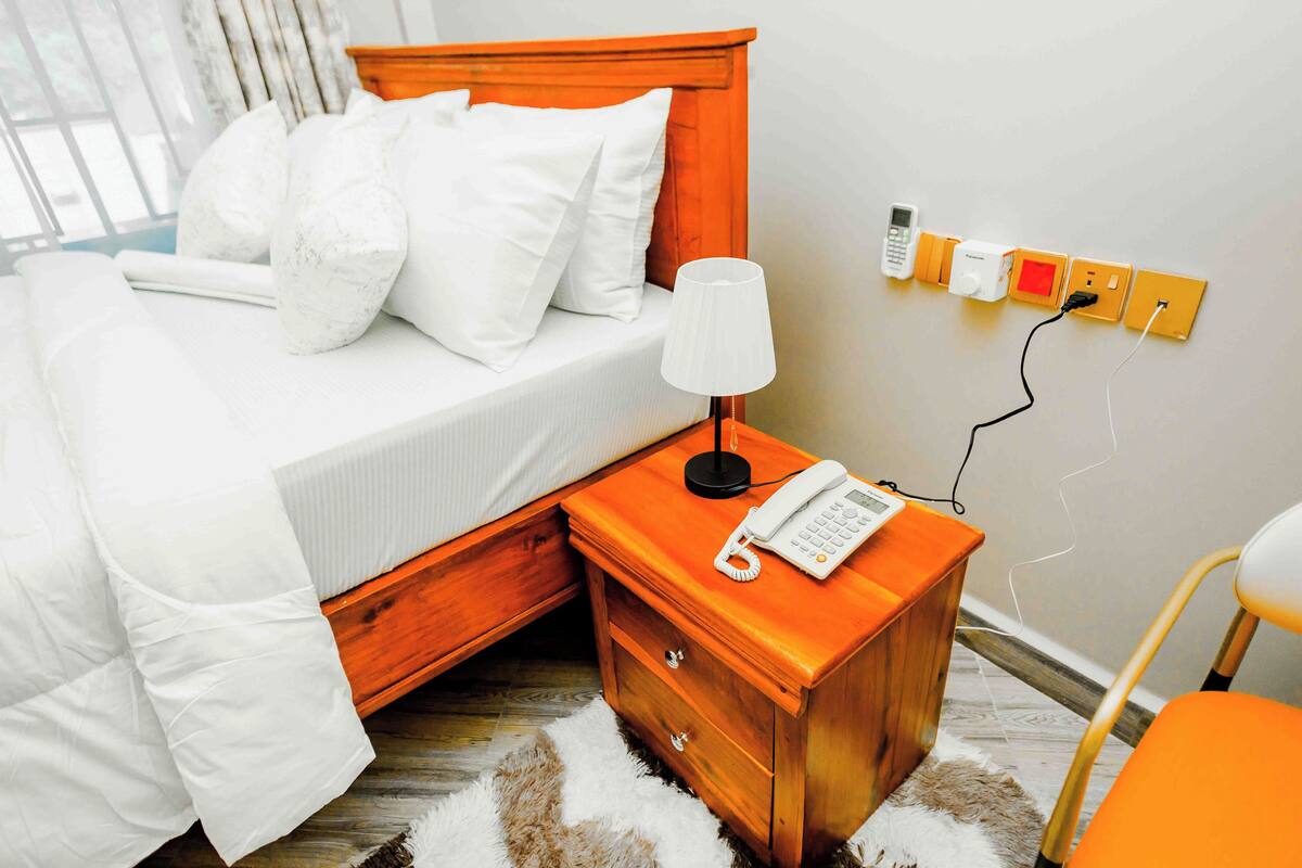 Una cama bien hecha con sábanas y almohadones blancos, una mesita de noche con lámpara y teléfono, y un enchufe mural con varios conectores.