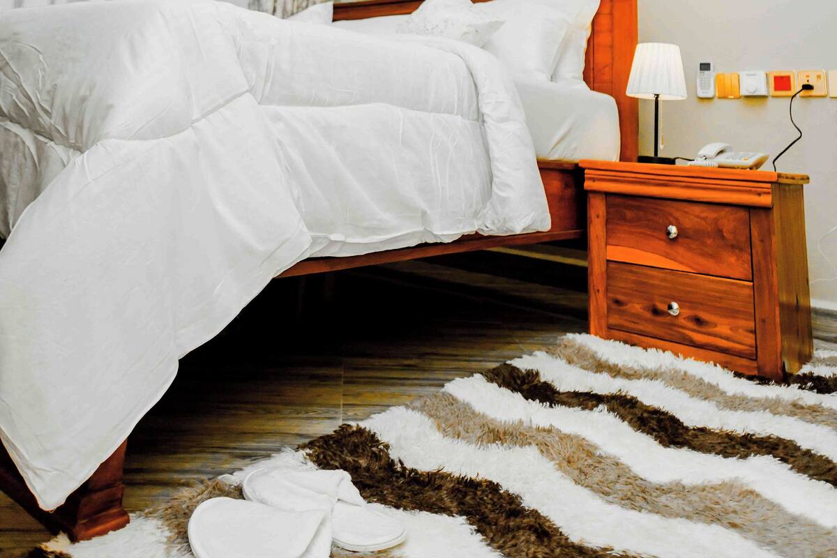 Una cama bien hecha con ropa de cama blanca, una mesita de noche de madera con lámpara, y una alfombra estampada en el suelo.