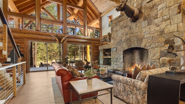 A spacious living room with a stone fireplace, wooden beams, and a large window overlooking a forest.