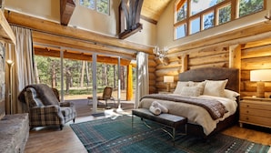 A log cabin bedroom with a large bed, a chair, and a view of the forest through large windows.