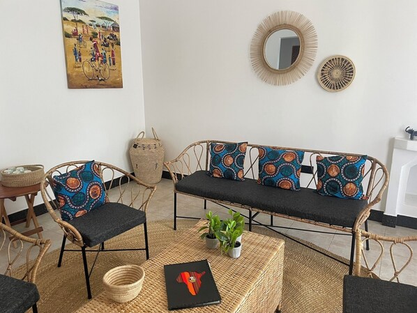 A room with a wicker sofa, two matching chairs, a woven basket, a small table with a bowl, and a framed painting on the wall. - Tanzania Safari Lodge (Arusha)