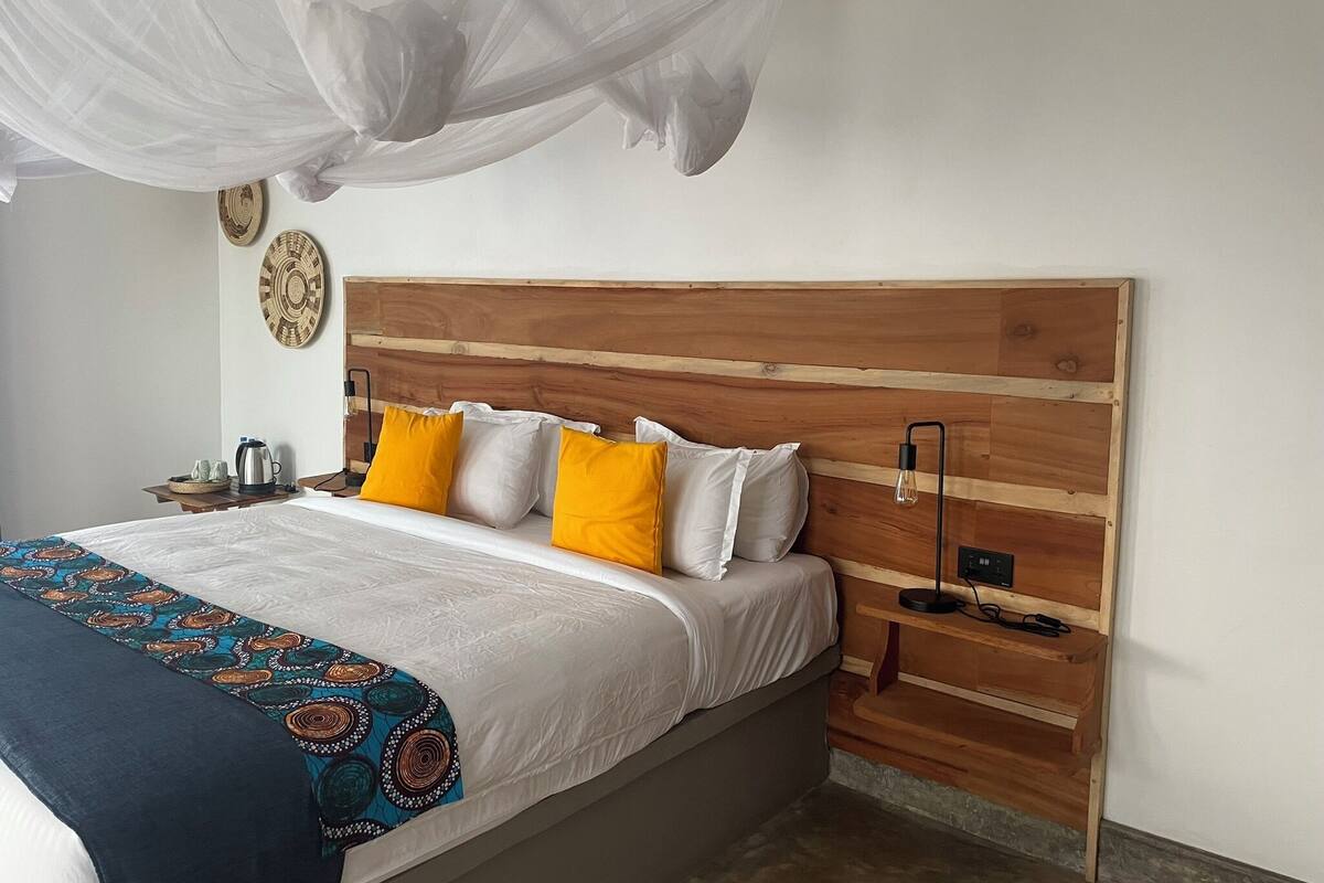 A bedroom with a wooden headboard, a bed with white and yellow pillows, a bedside table with a lamp, and a decorative wall hanging.