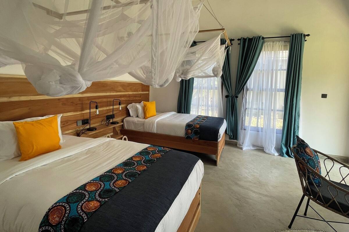 A hotel room with two beds, a canopy bed, a desk with a chair, and a view of the outdoors through large windows.