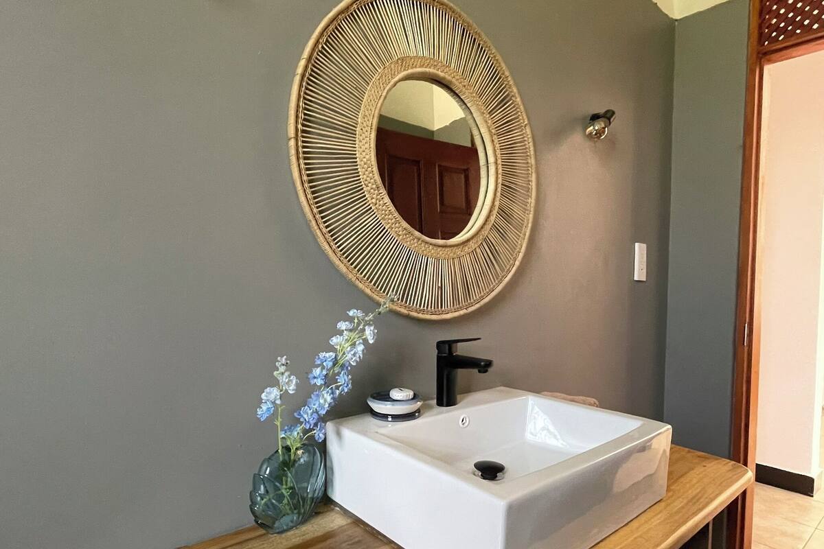 A bathroom with a white sink, a round mirror with a woven frame, and a vase with flowers.