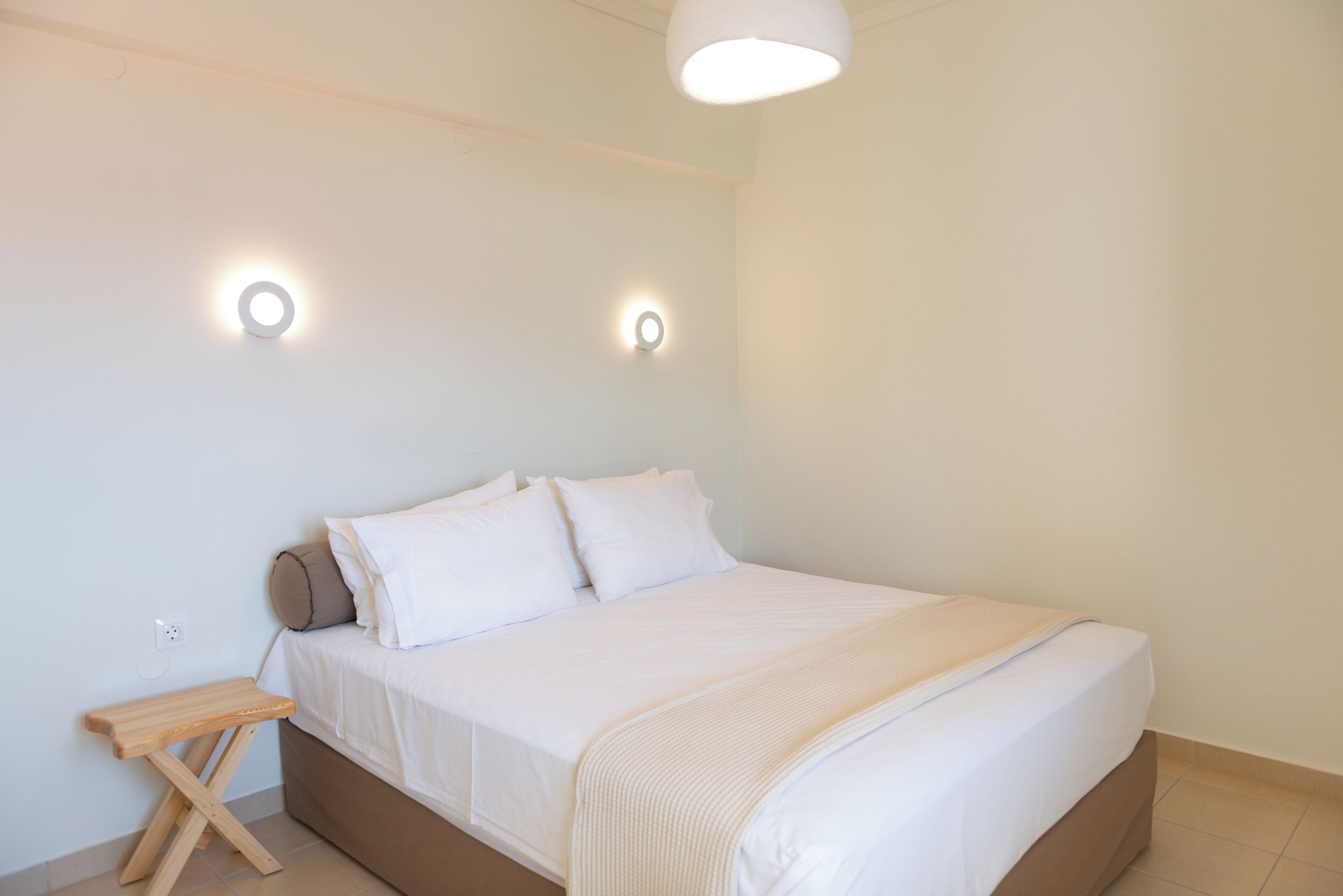 Un letto rifatto con lenzuola bianche, un comodino in legno e due lampade fissate al muro.
