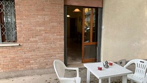 Uma área de pátio com uma mesa branca e duas cadeiras brancas, uma parede de tijolos e uma porta de vidro deslizante que dá para o interior.