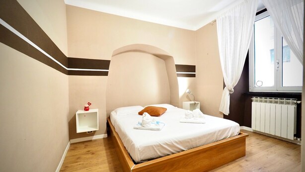 A bedroom with a wooden bed, a nightstand, a bedside table, a radiator, and a window with curtains. - Dimora delle Camelie (Aquarium) genovabnb it (GENOVA)