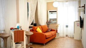 A living room with a sofa, a coffee table, a bar stool, and a small kitchen area with a refrigerator and a sink. - Dimora delle Camelie (Aquarium) genovabnb it (GENOVA)