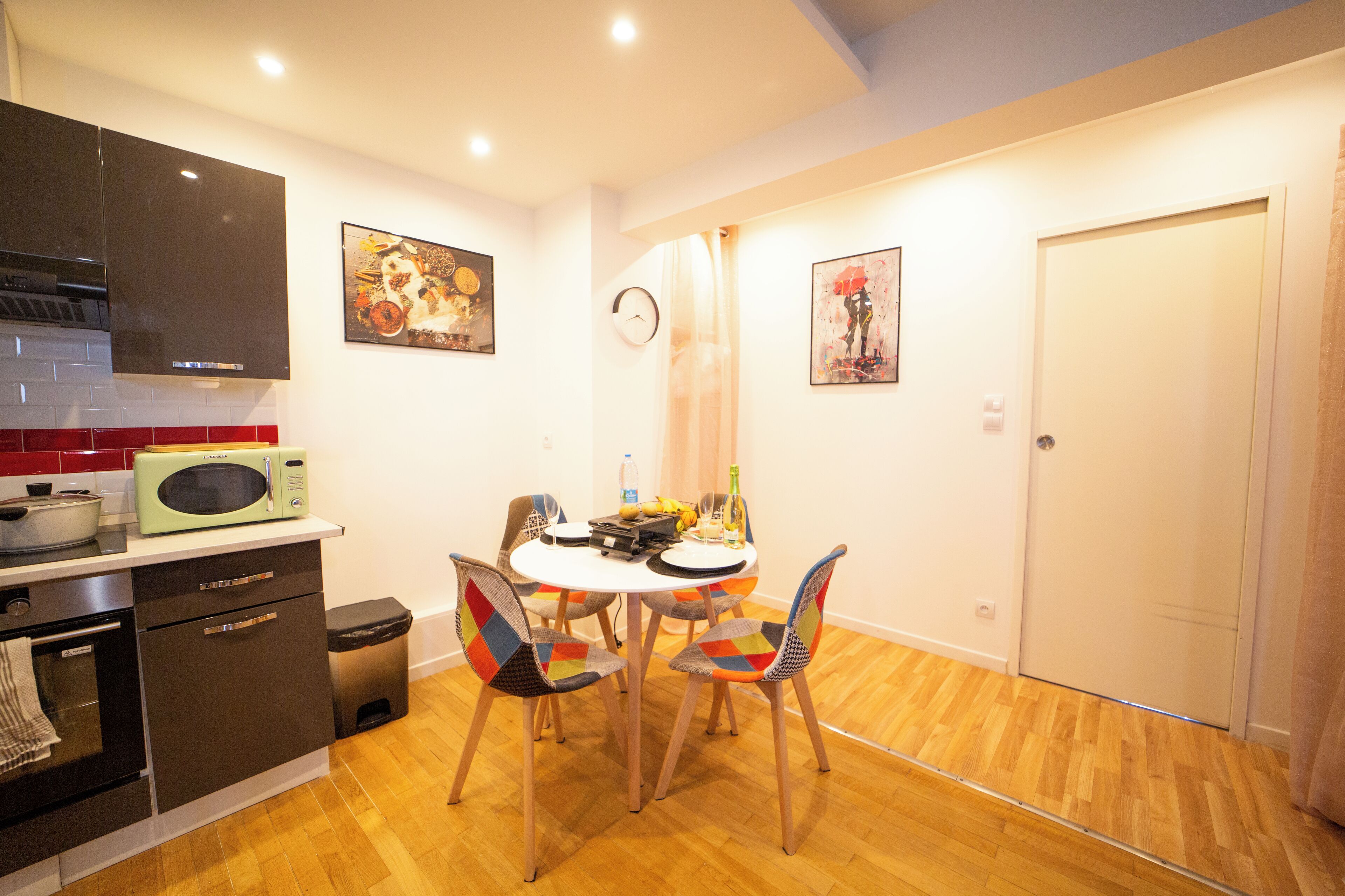 Cocina compacta con microondas, estufa y armarios. Hay una mesa de comedor con sillas y un reloj en la pared.
