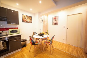 Cocina compacta con microondas, estufa y armarios. Hay una mesa de comedor con sillas y un reloj en la pared.