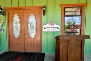 Een dubbele houten deur met een ruit, een bord met de tekst 'Wild Women Saloon Bar & Grill' en een houten bankje buiten.