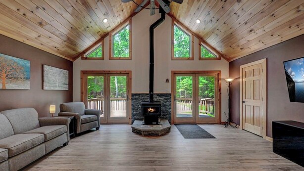 A cozy living room with a stone fireplace, wooden ceiling, and large windows. - NEW 4-bedroom home, private, hot tub, fenced yard, pet friendly (Hawley)