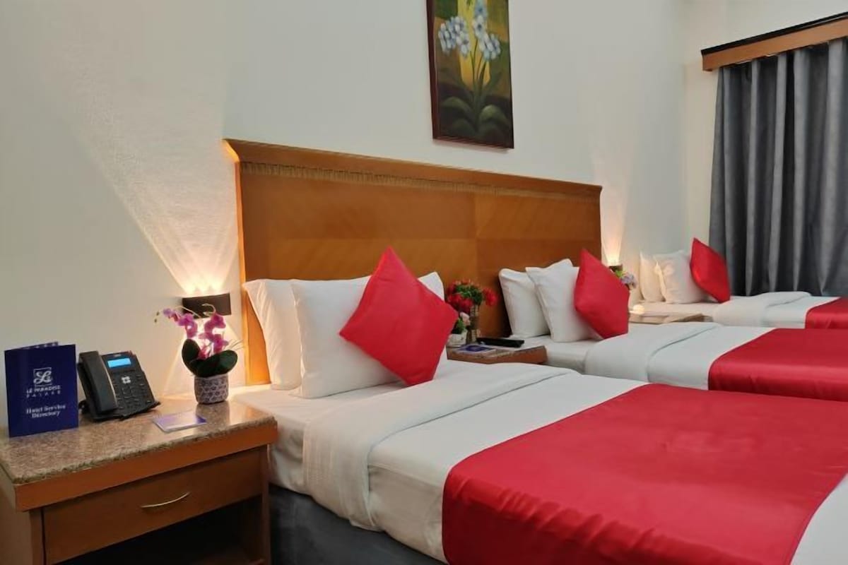 Habitación de hotel con dos camas, cada una con ropa de cama roja y blanca, cabecero de madera, mesita de noche con un teléfono y un letrero azul, además de un cuadro en la pared.