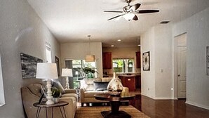 A living room with a ceiling fan, a sofa, a coffee table, and a rug. There is a kitchen in the background with cabinets and a window. - Charming 2-bedroom townhome in beautiful Riverview with WiFi, AC (Riverview)