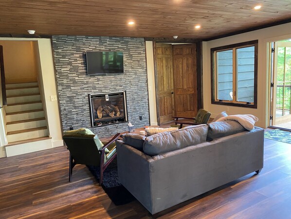 A modern living room with a stone fireplace, a large grey sofa, and a wooden staircase.