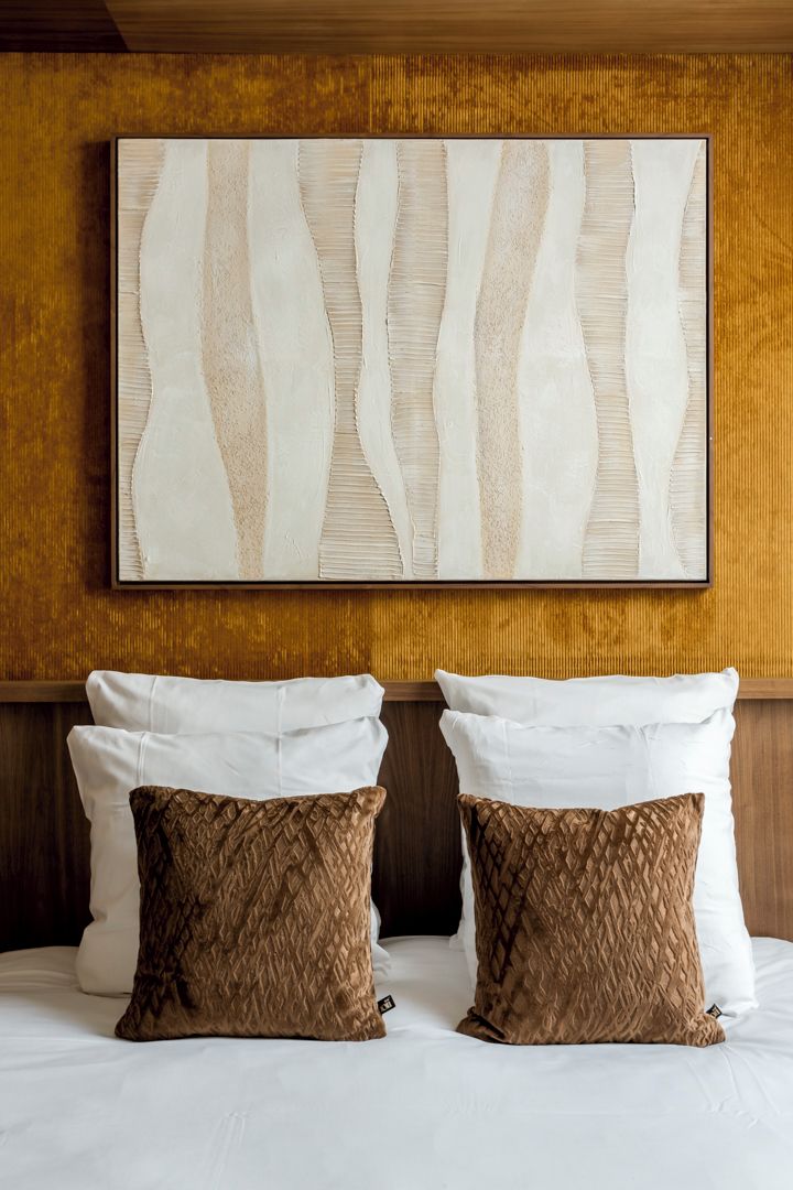 A hotel room with a large abstract painting on the wall, two pillows on the bed, and a wooden headboard.