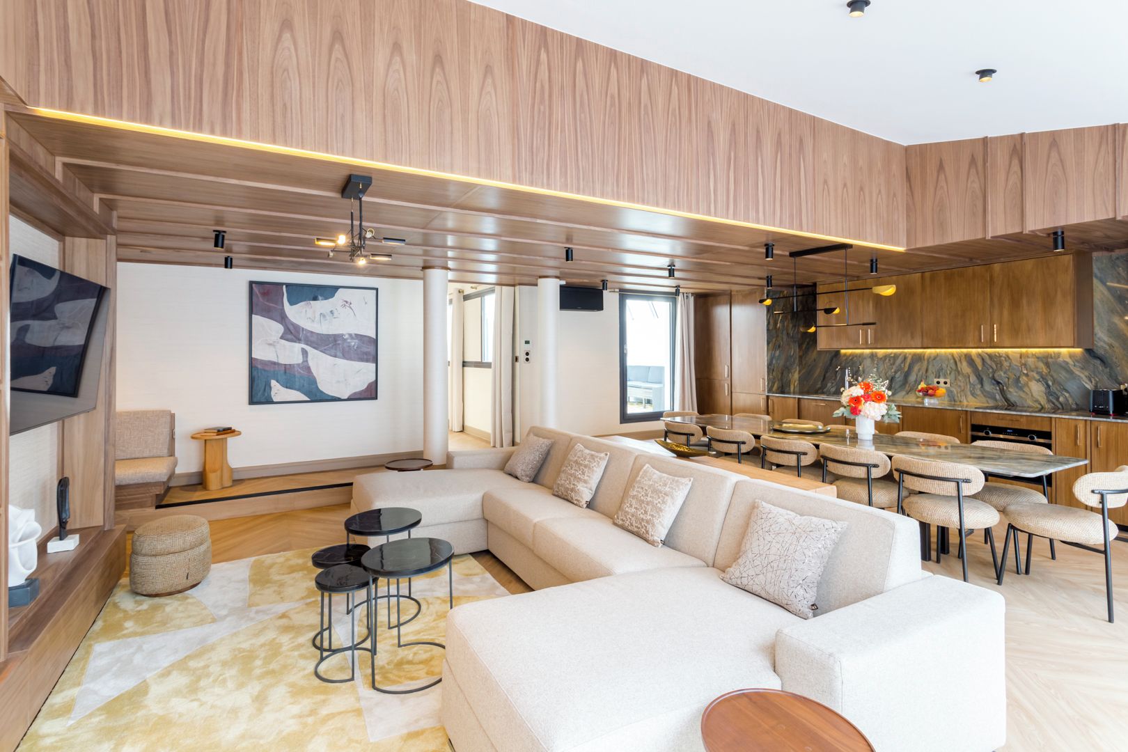 A modern living room with a large sectional sofa, a dining area with round tables, and a kitchen with wooden cabinets and a marble countertop.