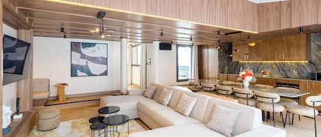 A modern living room with a large sectional sofa, a dining area with round tables, and a kitchen with wooden cabinets and a marble countertop.