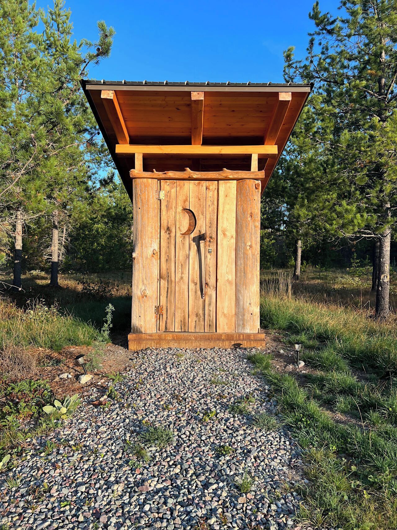 Et træskur med skråt tag, en dør med en halvmåneudskæring, og en grusvej, der fører hen til det.