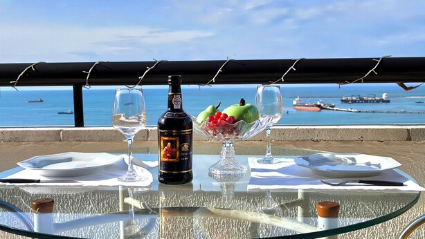 A table with a bottle of Dom Jose Ruby wine, two glasses, and a fruit bowl with green apples and red berries, set against a coastal view. - Flat Frente Mar com Terraço, perto Gran Marquise hotel  e praia (Meireles Fortaleza)