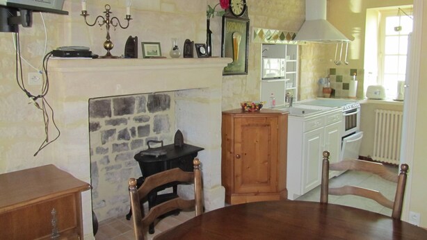 A cozy kitchen with a stone wall, wooden beams, a fireplace, and a dining area with a table and chairs.