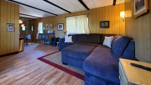 A cozy living room with a blue sofa, wooden walls, a red rug, and a dining area in the background.