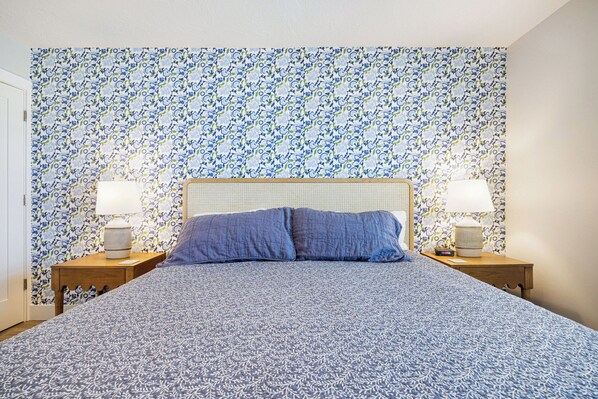 A bedroom with a patterned wallpaper, a bed with a blue and white bedspread, two bedside tables with lamps, and a wooden headboard. - Ocean's Reach 1A4, Direct Gulf-Front, All Amenities Open, Top Floor/Corner (Sanibel Island)