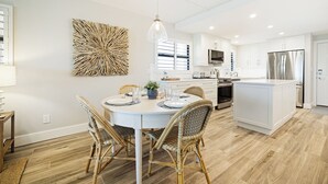 Een moderne keuken met witte kasten, een centraal eiland en een eethoek met rieten stoelen en een ronde tafel.